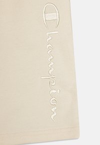 Beigts audums ar gludu tekstūru, uz kura ir izšūta "Champion" logo off-white krāsā, novietota vertikāli gar malām.