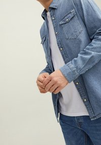 Camisa de mezclilla azul clara con dos bolsillos en el pecho, parte delantera abotonada y mangas largas, superpuesta a una camiseta blanca y combinada con jeans azules.
