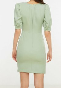 Robe verte ajustée avec des manches courtes bouffantes, une fermeture éclair dans le dos et des fronces sur les côtés. Tissu lisse avec un léger éclat.
