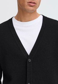 Black cardigan made of a textured knit, featuring a deep V-neck and three front buttons, worn over a white T-shirt.