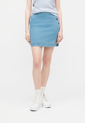 Donna che indossa una minigonna di denim azzurro con bottoni laterali, top a righe bianche, calzini bianchi e sneakers bianche con plateau, in piedi su sfondo bianco.
