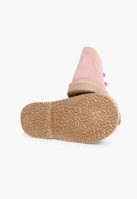 Bota de ante rosa con suela texturizada marrón visible, colocada en posición vertical sobre un fondo blanco, destacando el patrón de agarre debajo.