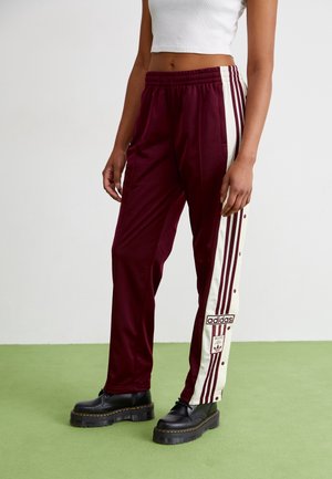 Osoba ubrana w bordowe spodnie dresowe Adidas z białymi paskami po bokach, czarne buty na platformie i biały krótki top, stoi na zielonej podłodze.