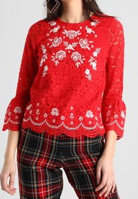 Top rosso in pizzo con ricami floreali, orlo scallop e maniche a campana a 3/4, abbinato a pantaloni a quadri con una combinazione di rosso, nero e bianco.