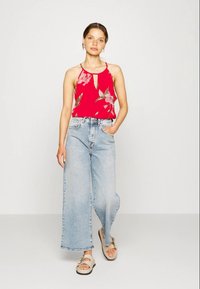 Top de alças vermelha com padrão floral e detalhe em buraco, combinado com jeans de perna larga azul claro. O visual é finalizado com sandálias bege.