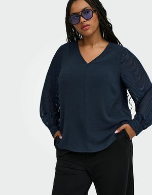 Donna con capelli intrecciati che indossa una camicetta blu scuro con maniche trasparenti a motivo, pantaloni neri e occhiali da sole con lenti azzurre, in piedi con le mani in tasca.
