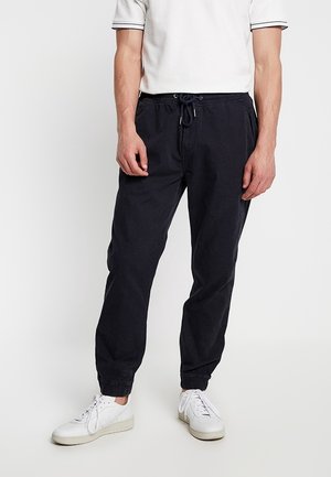 Homme portant un pantalon de jogging noir à cordon de serrage, des baskets blanches et un polo blanc à manches courtes, se tenant contre un fond uni.