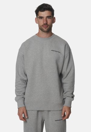 Man met kort donker haar die een bijpassende grijze trui en joggingbroek draagt met kleine zwarte tekst op de borst en dij.