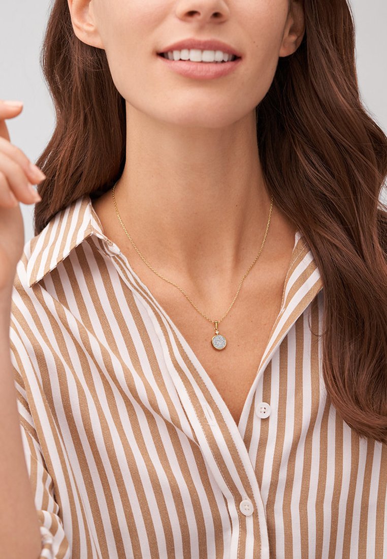 Złoty naszyjnik z łańcuszkiem, który ozdobiony jest okrągłym, fakturowanym zawieszką. Model nosi koszulę w paski z guzikami w jasnobrązowe i białe paski.
