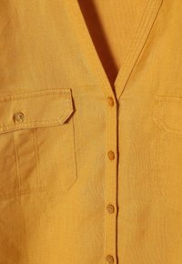 Camicia di lino gialla con scollatura a V, due tasche sul petto e chiusura a bottoni. Texture liscia con dettagli di cucitura discreti.
