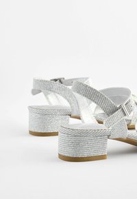 Zilveren sandalen met hak hebben gestructureerde banden en een blokhak, met een gladde zool. Het ontwerp bevat een gespsluiting en een glinsterende afwerking.