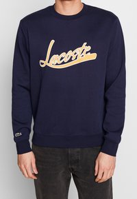 Mörkblå sweatshirt i bomull med en stor beige "Lacoste"-logotyp, ribbade ärmslut och nederkant, samt en grön logotypaccent på ärmen.