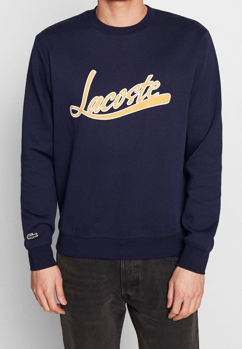 Mörkblå sweatshirt i bomull med en stor beige "Lacoste"-logotyp, ribbade ärmslut och nederkant, samt en grön logotypaccent på ärmen.