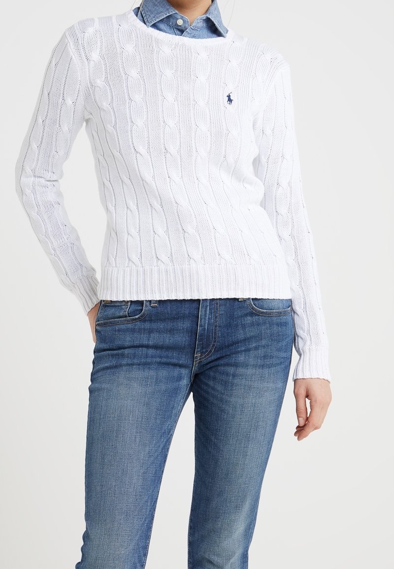 Femme portant un pull blanc en maille torsadée par-dessus une chemise en denim bleu et un jean bleu, debout avec une main dans la poche sur un fond uni.
