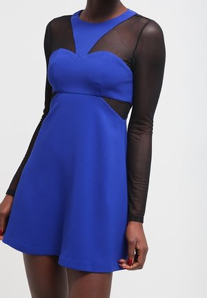 Femme portant une robe bleue sans manches avec de longues manches noires transparentes et des découpes sur les côtés sous la poitrine.