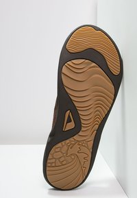 Brun atletisk skosåle med teksturert gummimønster; har bølgete linjer og en småsteinlignende design; mørk øverste del med brune detaljer.