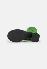 Bottine en daim vert avec un talon épais noir et une pointe arrondie lisse. La vue inférieure montre une semelle noire solide.