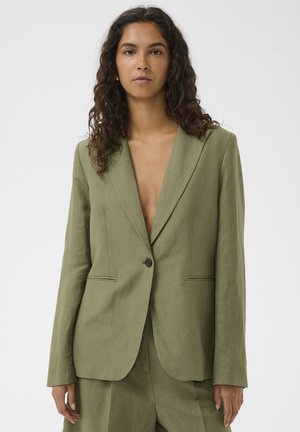 Naine lokkis tume juustega, kandes lahtist oliivrohelist bleiserit ja sobivaid pükse ühtlase tausta ees.