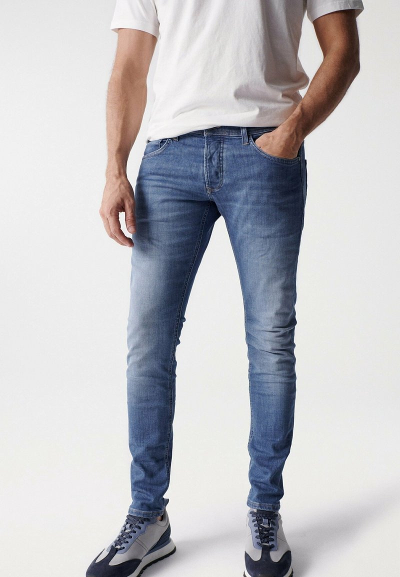 Homme portant un jean slim bleu, un t-shirt blanc et des baskets gris-bleu, debout avec une main dans la poche sur un fond uni.