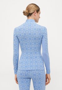 Kvinne med blondt hår i en knute iført en lys blå mønstret langermet topp og matchende leggings, vist fra ryggen på en hvit bakgrunn.