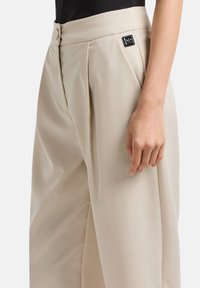 Pantaloni beige su misura con pieghe, dotati di chiusura a bottone e una piccola etichetta nera sul cinturino. Tessuto dalla consistenza liscia.