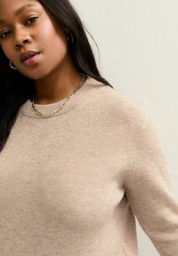 Beżowy sweter z dzianiny z okrągłym dekoltem, miękka tekstura i delikatne fleki. Złoty łańcuszek jako akcent. Włosy ułożone w luźne fale.