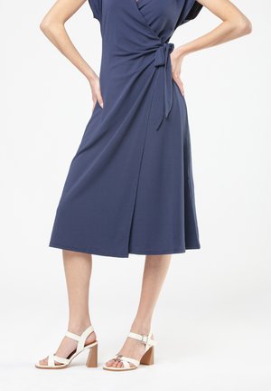 Vrouw in een marineblauwe midi-jurk met wikkelontwerp, gekleed in witte sandalen met blokhak en bandjes, staande met haar handen op haar heupen tegen een witte achtergrond.