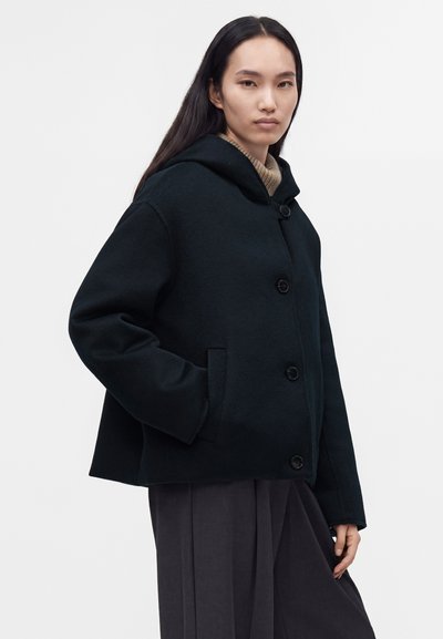 Femme aux longs cheveux foncés portant un manteau noir à capuche boutonné et un pantalon plissé gris, debout devant un fond clair uni.
