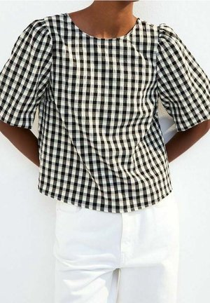 Vrouw met een zwart-wit geruit kort mouwen topje en witte broek met hoge taille, staand met haar handen achter haar rug.