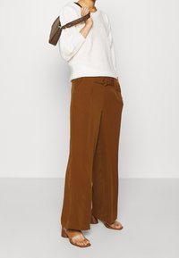 Pull blanc texturé, pantalon large marron avec un nœud à l'avant, et sandales marron à brides. Petit sac à main marron avec une bandoulière à motif.