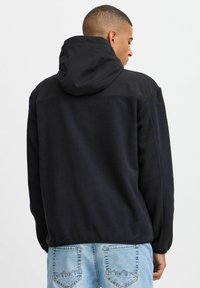 Svart fleecejakke med hette, med en glatt øvre del og en teksturert nedre del. Brukt sammen med lyseblå denimjeans.