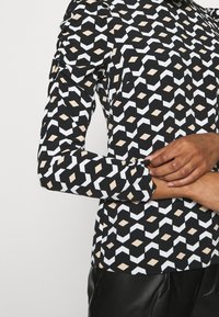 Černobílý top se geometrickým vzorem a krémovými akcenty, dlouhými rukávy a nabíranými detaily na ramenou, vyrobený z měkkého materiálu.