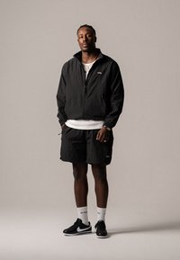 Giacca leggera nera con zip, pantaloni corti neri, T-shirt bianca, sneakers nere con dettagli bianchi, calzini bianchi con loghi, in posizione eretta.