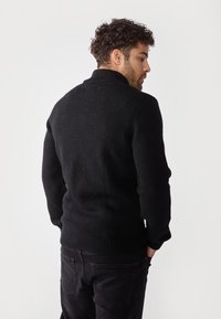 Suéter negro de punto con alto cuello, puños de canalé y dobladillo, combinado con jeans negros. La textura parece suave y ligeramente difusa.
