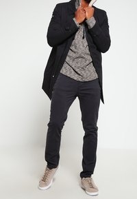 Musta pitkä takki harmaan teksturoidun hupparin päällä, yhdistettynä tummiin housuihin ja vaalean beigeihin lenkkikenkiin. Takissa on kauluksellinen kaula ja napit.