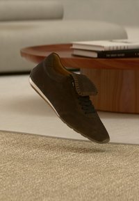 Σκούρο καφέ σουέντ αθλητικό παπούτσι με μαύρα κορδόνια και διπλωμένο φουλάρι στην κορυφή, αιωρούμενο πάνω από μπεζ χαλί σε ένα σαλόνι.