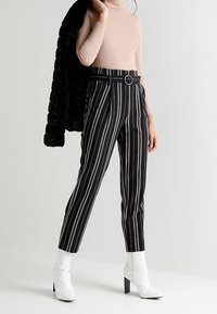 Svart och vitrandiga byxor med hög midja och en rund bältesknäppning, kombinerat med en åtsittande rosa polotröja och vita ankle boots.