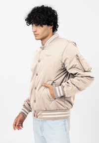 Beżowa bomberka wykonana z materiału przypominającego satynę, z zamkiem z przodu, ściągaczami przy mankietach oraz paskiem w paski, z boczną kieszenią i haftowanym logo.