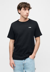 Νεαρός άνδρας με καστανά σγουρά μαλλιά, που φοράει μαύρο μπλουζάκι με την επιγραφή "CONS" και ανοιχτόχρωμο μπλε τζιν, στέκεται με το ένα χέρι στην τσέπη, κοιτάζοντας προς τα δεξιά.