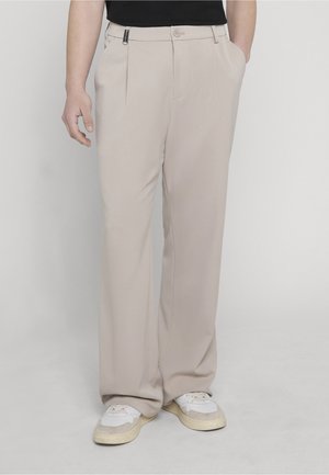 Persoană purtând pantaloni largi bej și adidași albi, stând cu mâinile în buzunare pe un fundal simplu.