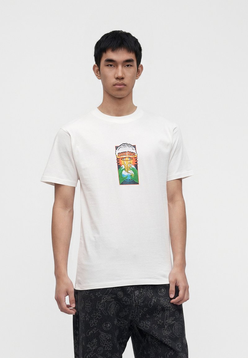 Νέος άνδρας φοράει λευκό T-shirt με πολύχρωμη γραφική απεικόνιση ενός πουλιού και κυμάτων, συνδυασμένο με μαύρο παντελόνι με σχέδια λουλουδιών και χαμογελαστών προσώπων.