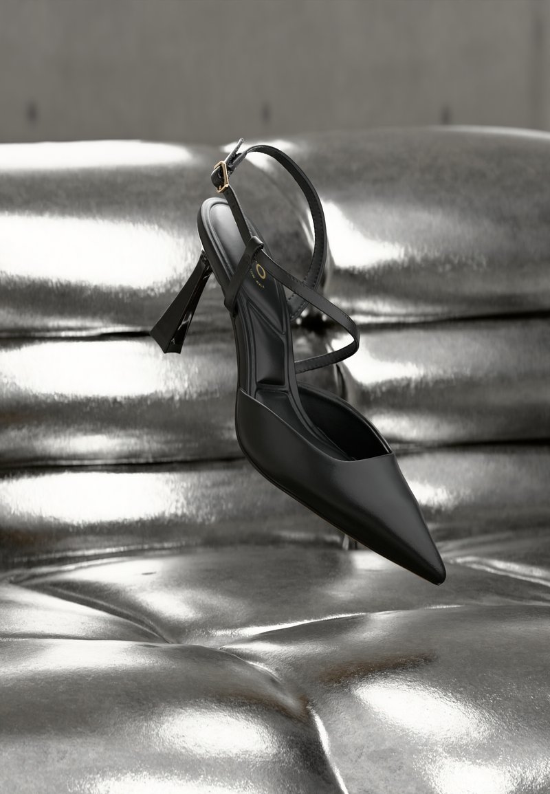 Chaussure à talon noir à bout pointu et dos ouvert. Comprend une fine bride autour de la cheville et une semelle texturée. Design épuré et minimaliste mis en valeur par un arrière-plan métallique brillant.