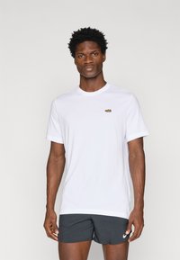 Bela kratka majica s kratkimi rokavi iz bombaža, ki ima majhen rjav logotip na prsih. Oblečena z temnimi kratkimi hlačami, preprostega dizajna.