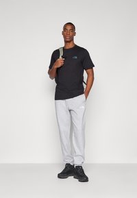 Camiseta negra con logo, pantalones de chándal grises y zapatillas negras. El modelo sostiene una mochila. Diseño simple, ajuste casual, sin patrones visibles.