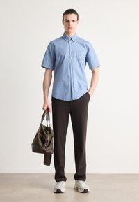 Lichtblauwe kraagloze short-sleeve blouse met een borstzak, gecombineerd met zwarte broek en een groene sporttas. Sportieve grijze sneakers.