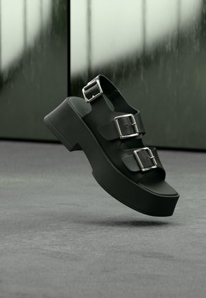 Μαύρο σανδάλι πλατφόρμα με τρεις φαρδιές λουριά και ασημένιες αγκράφες, να αιωρείται πάνω από γκρι χαλί με θολό γυάλινο φόντο.