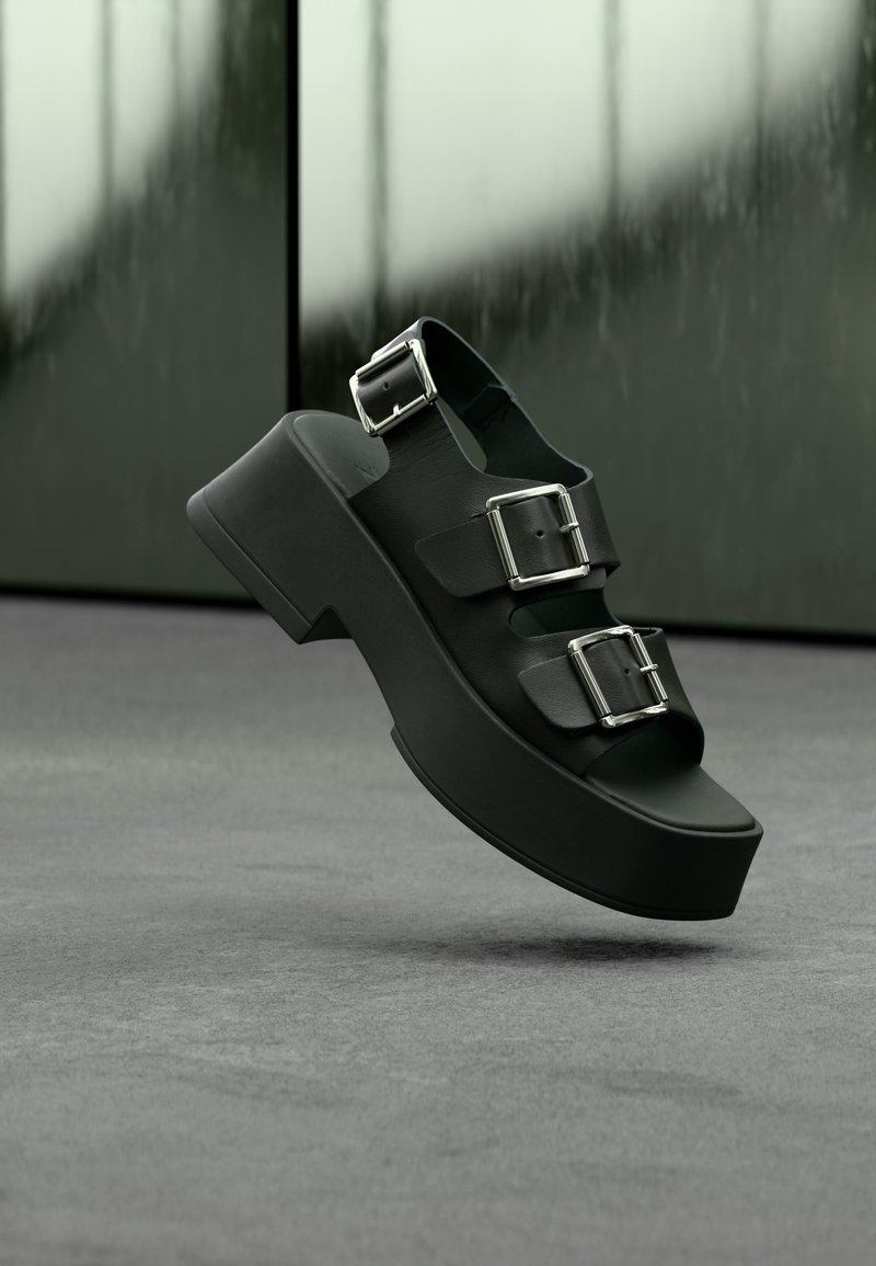 Μαύρο σανδάλι πλατφόρμα με τρεις φαρδιές λουριά και ασημένιες αγκράφες, να αιωρείται πάνω από γκρι χαλί με θολό γυάλινο φόντο.