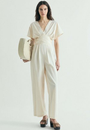 Mujer que lleva un mono crema estilo cruzado, sosteniendo un bolso grande tejido, combinado con sandalias marrones de plataforma, de pie frente a un fondo liso.