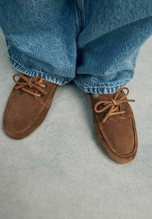 Scarpe mocassino in suede marrone con punta rotonda, chiusura con lacci e lacci a contrasto, abbinate a ampi jeans in denim blu.