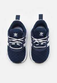 Pari tummansinisistä Adidas-taaperokenkiä valkoisilla nauhoilla ja ikonisilla kolmella raidalla, nähtynä ylhäältä valkoista taustaa vasten.